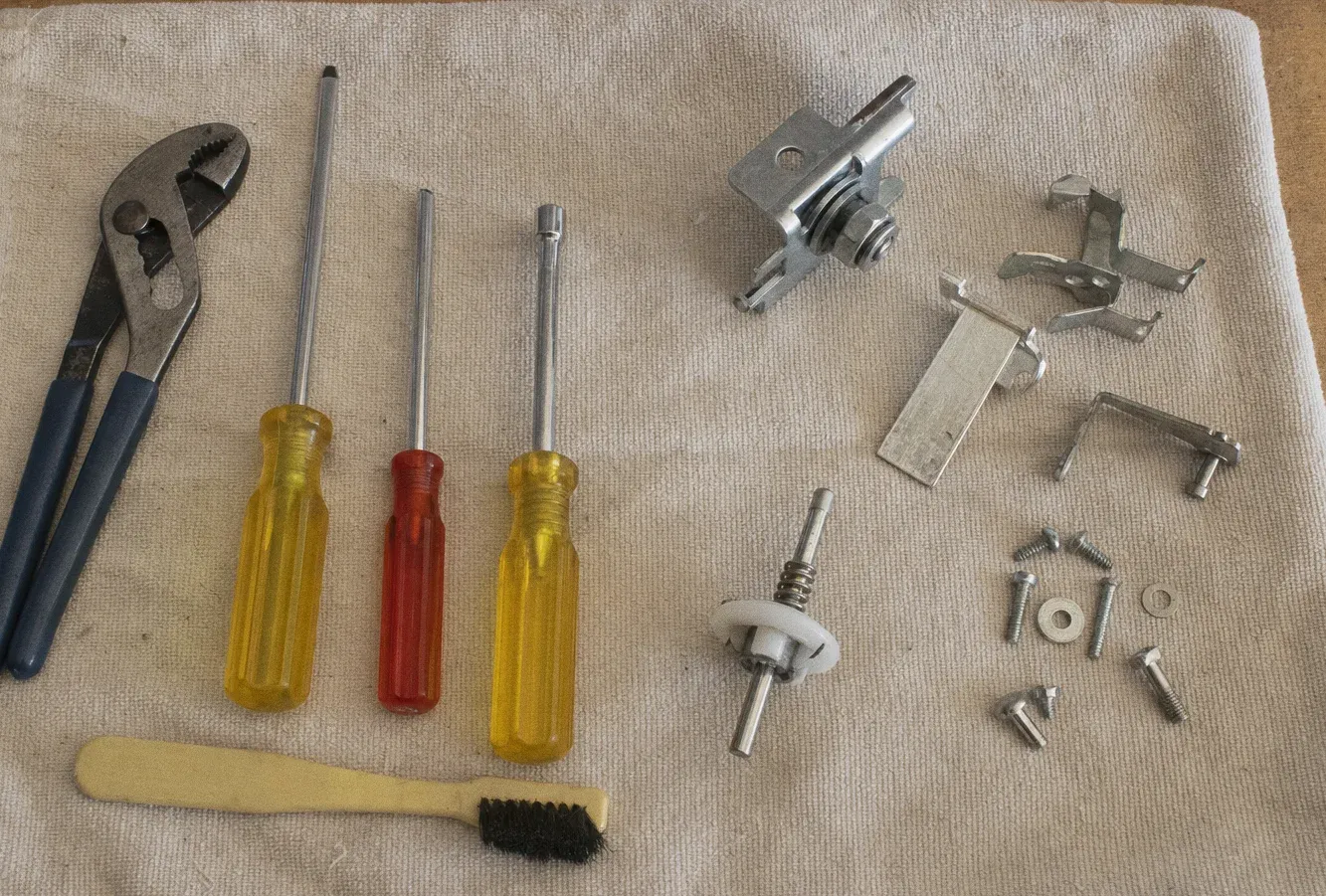 An adjustable pipe wrench, three nut drivers and a brush on a beige towel beside hinge brackets, a valve stem and small screws